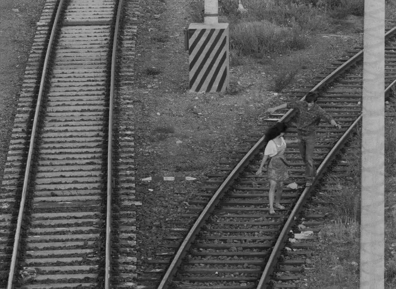 Pearls of the Deep (1965) movie still: wide — A pair of railway tracks runs through an open area with dirt and scattered weeds. A young man and…