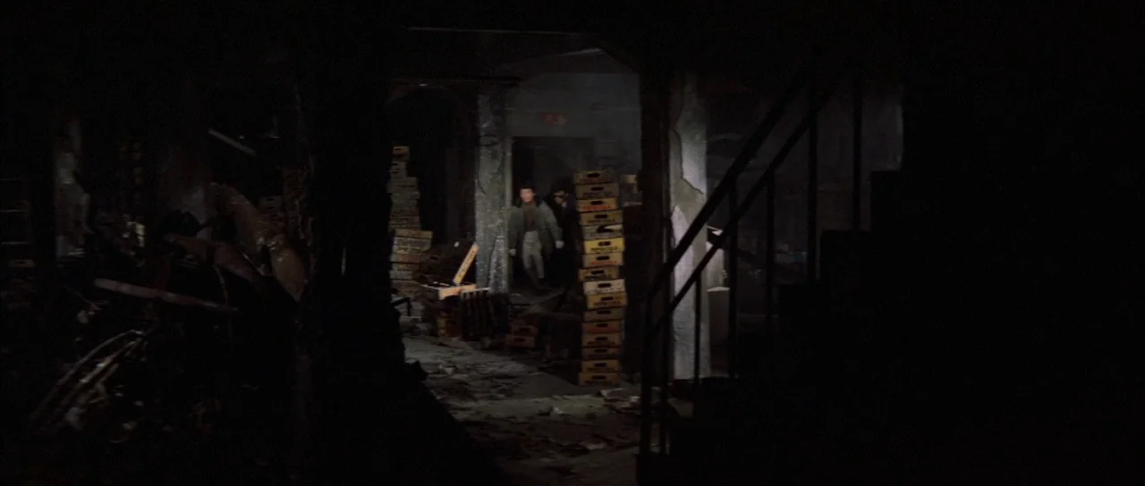 All Monsters Attack (1969) movie still: wide — A cluttered, dark basement filled with stacks of wooden crates and debris. Two figures walk away from…