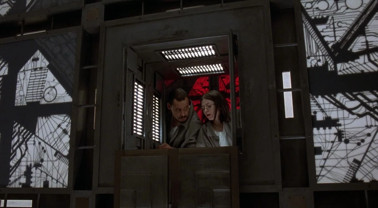 Cube (1997) movie still: medium — A dark, modular control room features metallic walls with glowing circuit board patterns. A man and woman stand…
