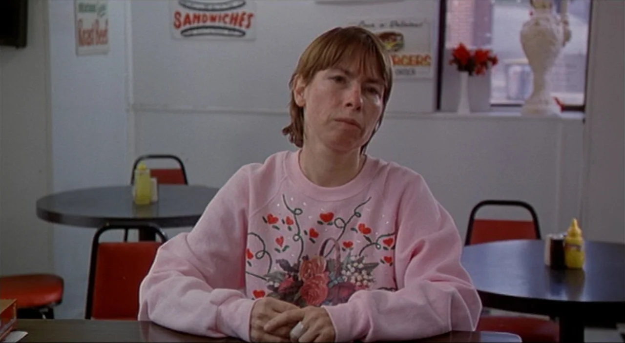 Pecker movie still: medium — A quiet diner interior featuring round tables and red vinyl chairs against white walls. A woman in a pink sweatshirt…