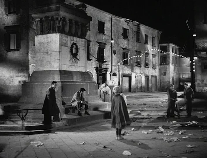 La Strada (1954) movie still: wide — An Italian-style piazza at night features a large stone monument and surrounding multi-story buildings. Litter…