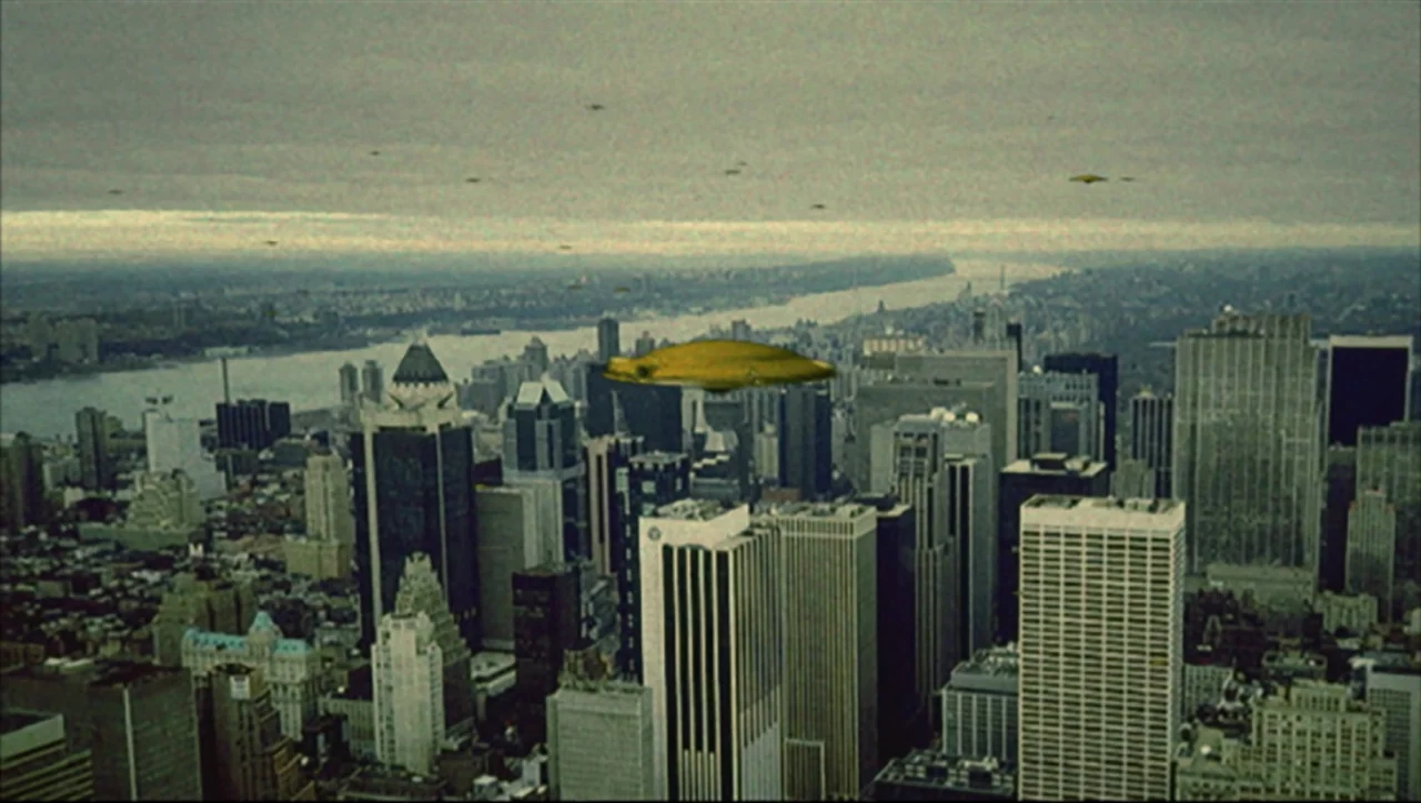Wild Zero (1999) movie still: establishing — A wide aerial view of the New York City skyline under a gray cloudy sky. Multiple golden saucer-shaped…