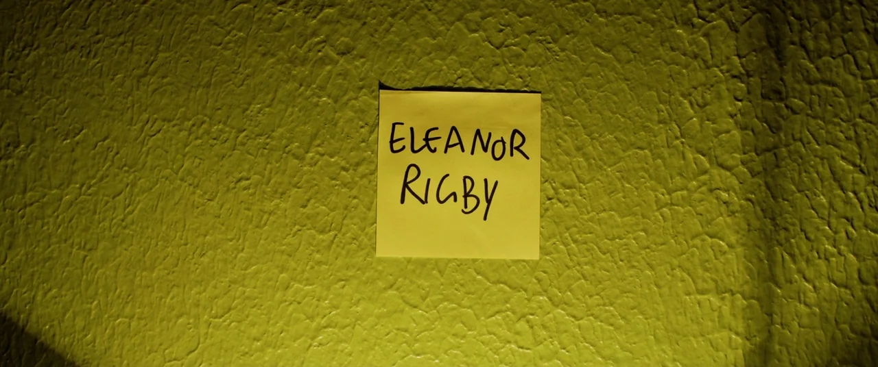Yesterday (2019) movie still: closeup — A bright yellow sticky note with handwritten text is centered on a textured wall. The textured wall is…