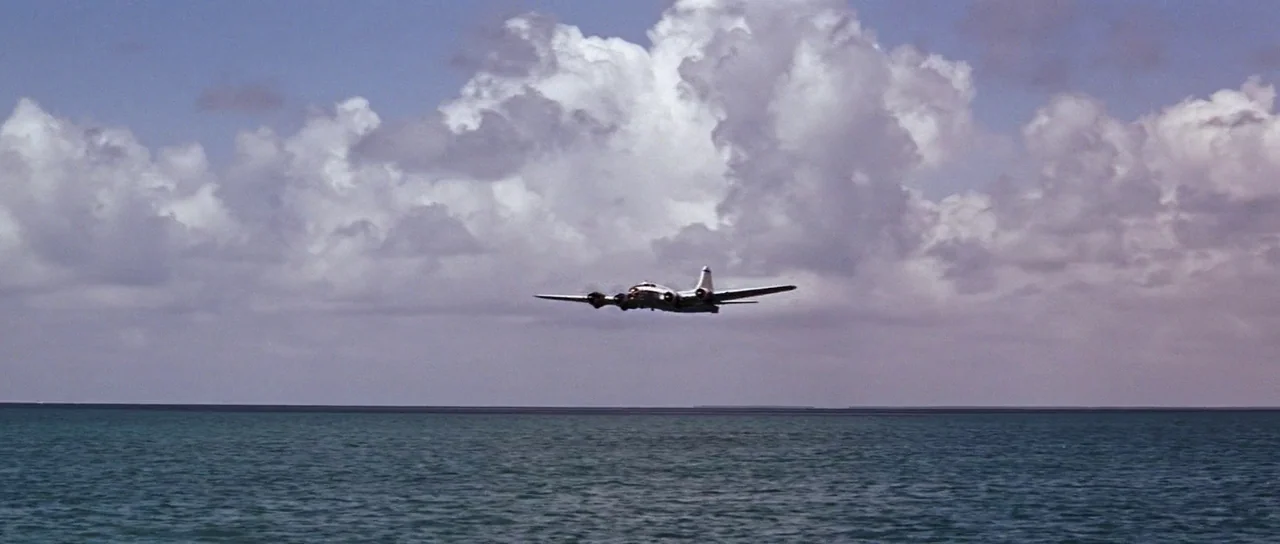 Thunderball (1965) movie still: wide — A silver multi-engine propeller aircraft flies low over calm, vast ocean waters. Large white cumulus clouds…