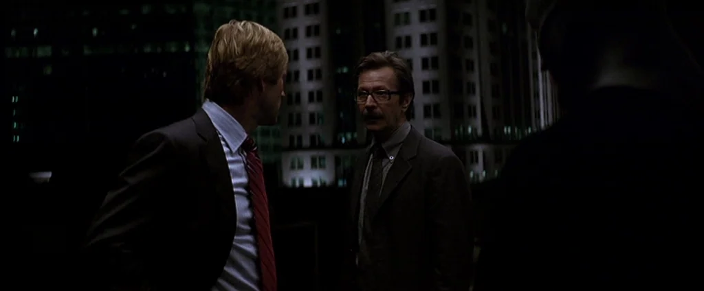 The Dark Knight (2008) movie still: medium — A dark outdoor terrace overlooks illuminated city high-rise buildings at night. Three men stand in…