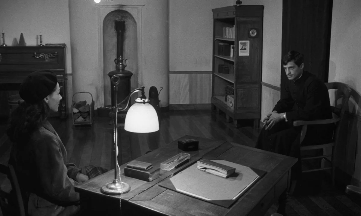Léon Morin, Priest (1961) movie still: medium — A sparsely furnished room features a central wooden desk with a lamp and books. A woman wearing a…