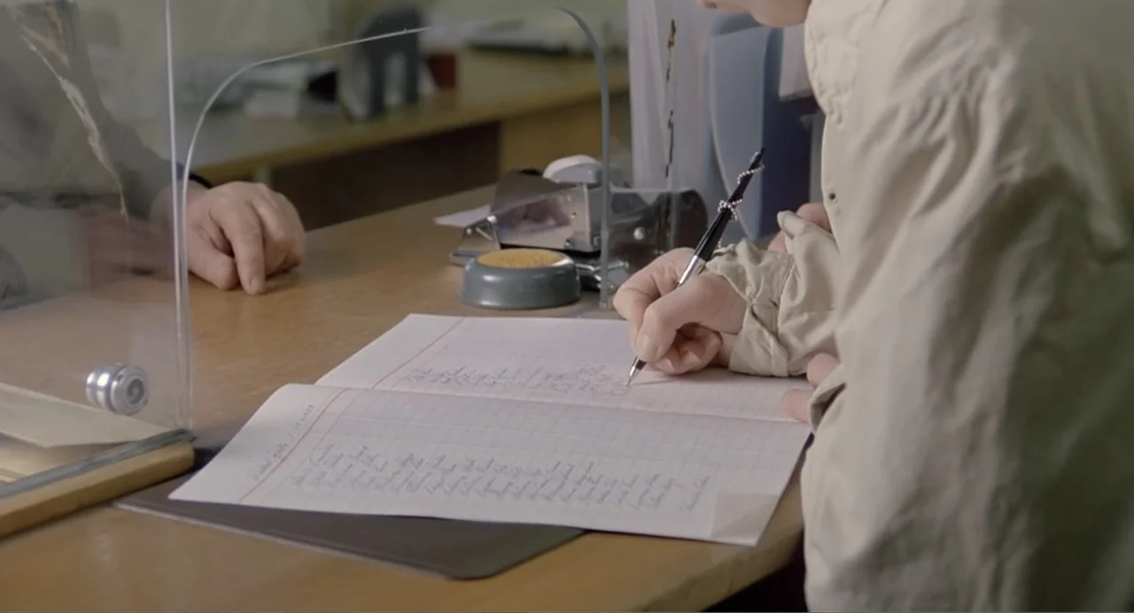 The Match Factory Girl (1990) movie still: medium — An administrative service counter features a glass partition and a ledger book. A person writes…