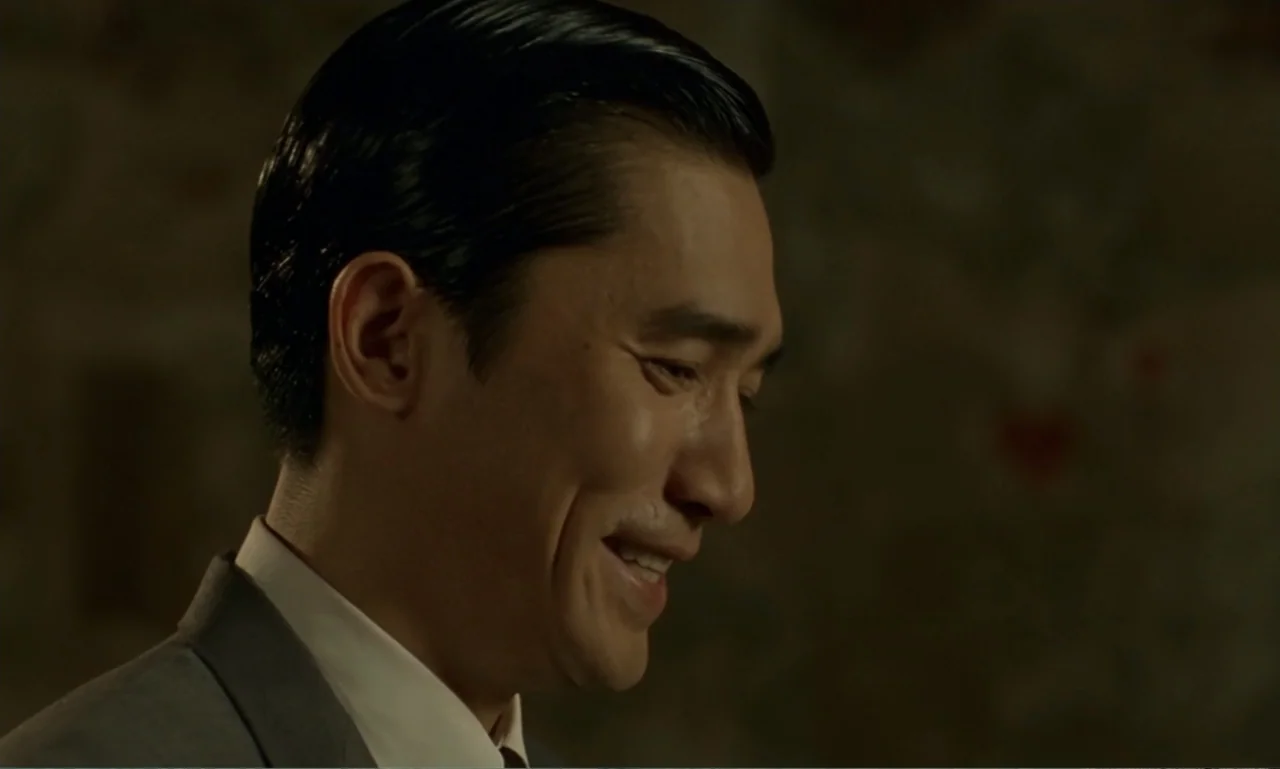 In the Mood for Love (2000) movie still: closeup — An interior space features a dimly lit office with neutral wall tones. A man in a grey suit…