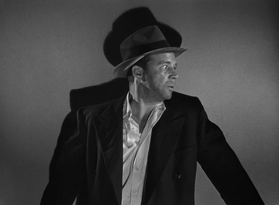 Murder, My Sweet (1944) movie still: medium — A man stands against a stark, featureless wall illuminated by high-contrast studio lighting. He wears…