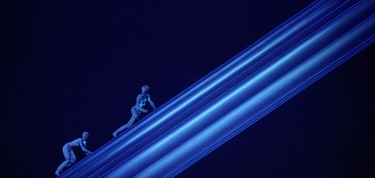 Tron (1982) movie still: wide — Two individuals climb a massive glowing blue diagonal beam set against a black void. The environment consists of…