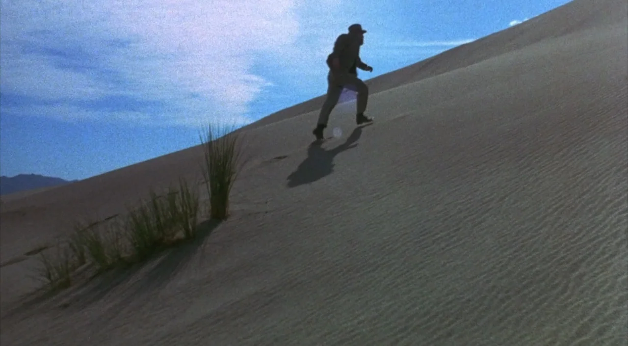 Dr. Phibes Rises Again (1972) movie still: wide — A vast expanse of undulating sand dunes stretches under a bright blue sky. A solitary man treks…