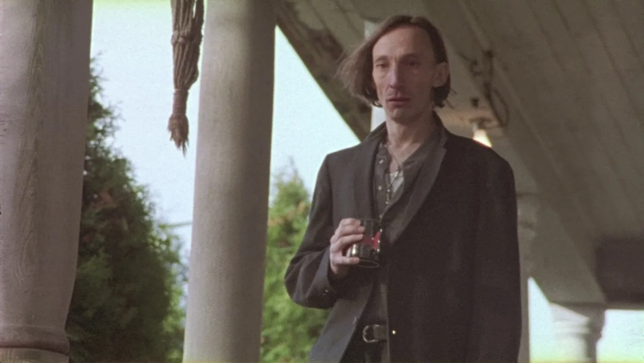 Hard Core Logo movie still: medium — A residential porch features round pillars and a hanging rope against a green background. An older thin man in…