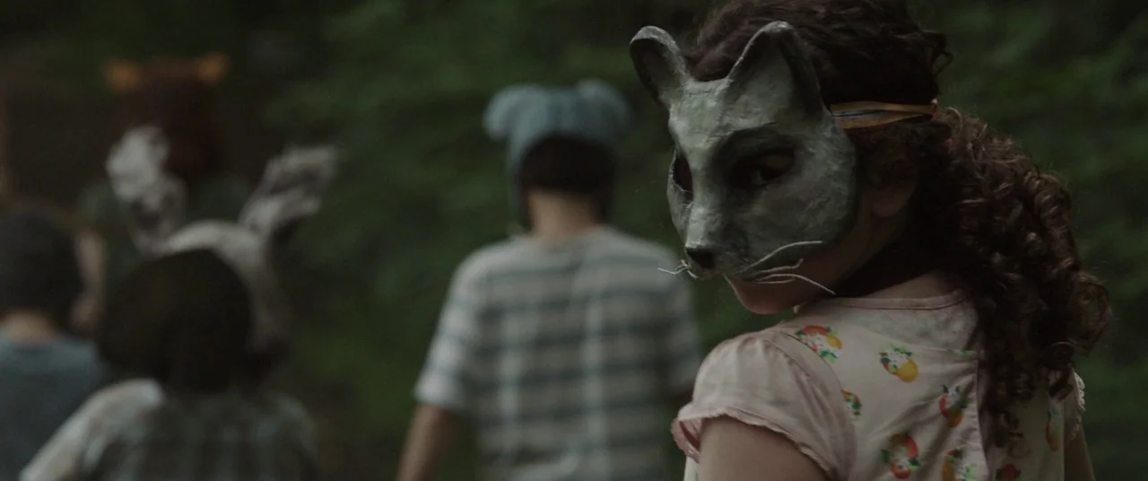 Pet Sematary (2019) movie still: medium — A group of children wearing animal masks walk through a dense, tree-covered outdoor space. One child in…