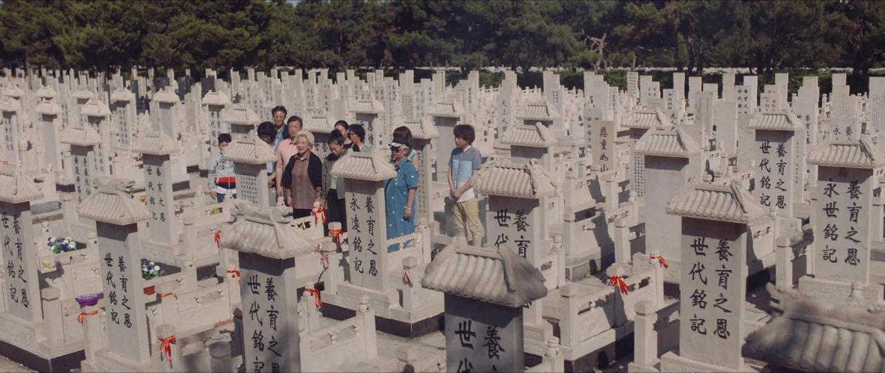 The Farewell (2019) movie still: wide — A dense cemetery features rows of light stone markers with traditional tiered roof structures. A small group…