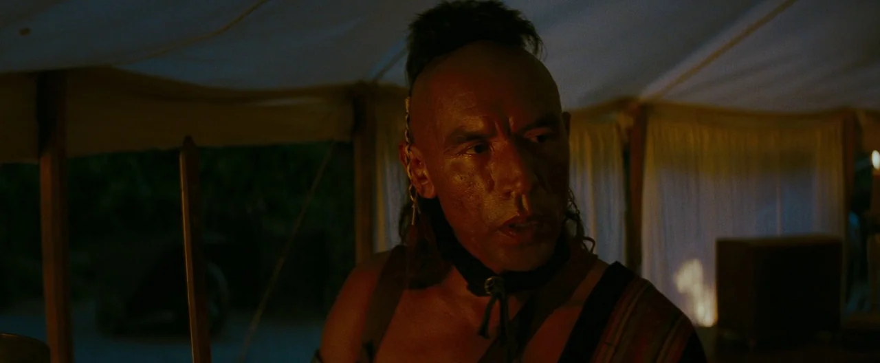 The Last of the Mohicans (1992) movie still: medium — Inside a dim canvas tent with draped walls, lit by flickering warm firelight. A man with a…