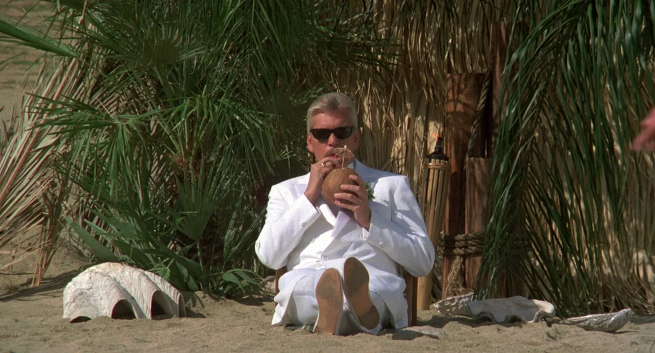 Night of the Creeps (1986) movie still: medium — A man sits on sand in front of palm fronds and a thatch screen. He is wearing a white suit and…