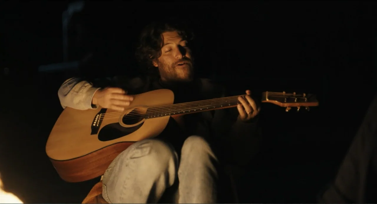Hell of a Summer (2025) movie still: medium — A dark outdoor setting at night features a man sitting near a campfire. The man plays a wooden…