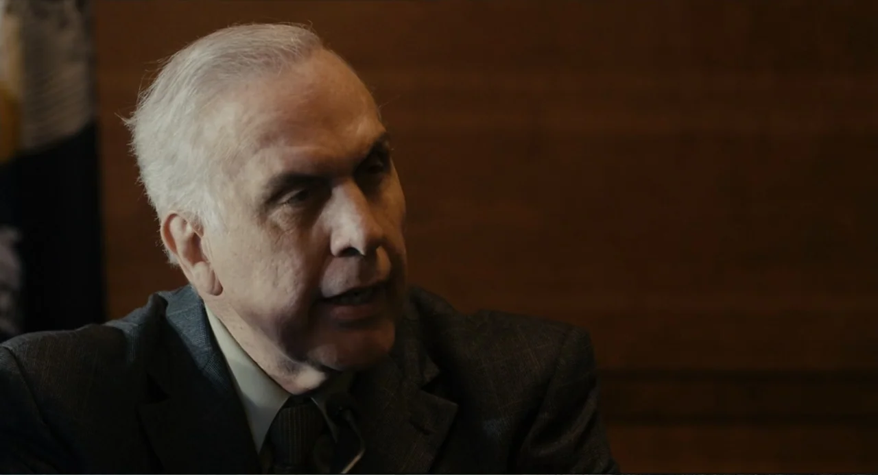 Beyond a Reasonable Doubt (2009) movie still: closeup — A dimly lit room with wood-paneled walls in the background. An elderly man in a suit speaks…