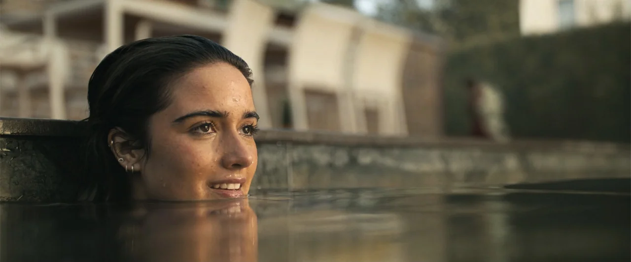 My Fault: London (2025) movie still: closeup — A woman rests her chin at the water level of a modern outdoor pool. She looks out from the water…