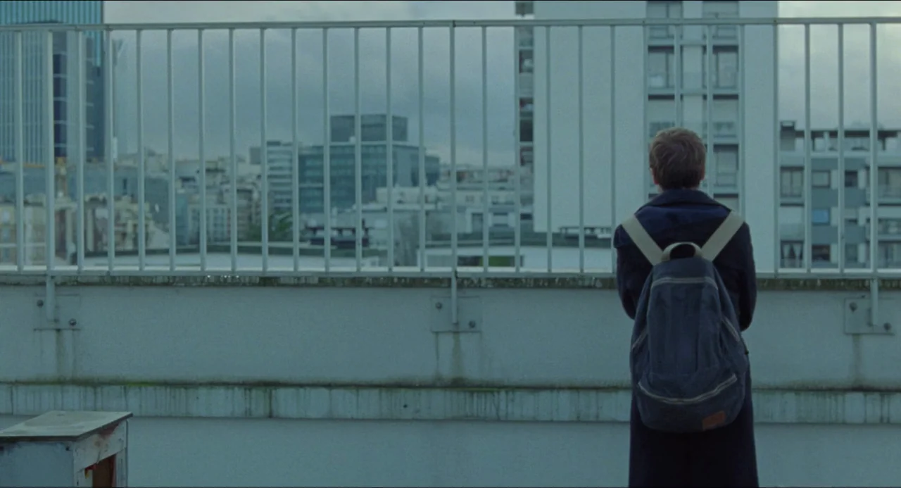 One Fine Morning (2022) movie still: fullbody — A concrete rooftop terrace overlooks an urban skyline of residential and office buildings. A young…