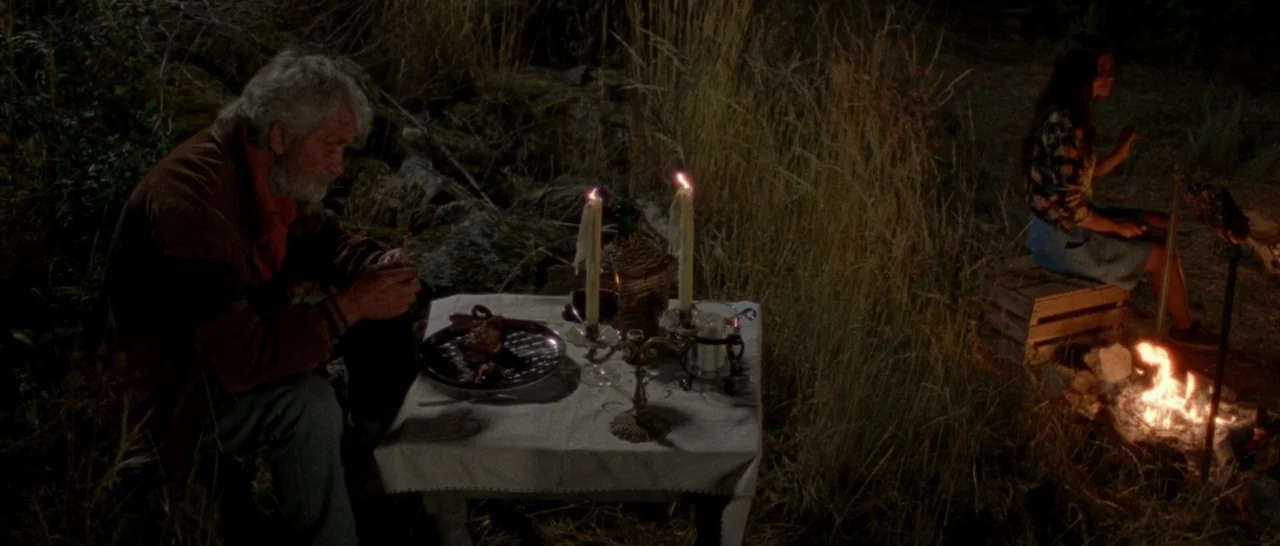 Arabian Nights: Volume 2 The Desolate One (2015) movie still: wide — An outdoor campsite at night with tall grass and a small table set for dinner.…
