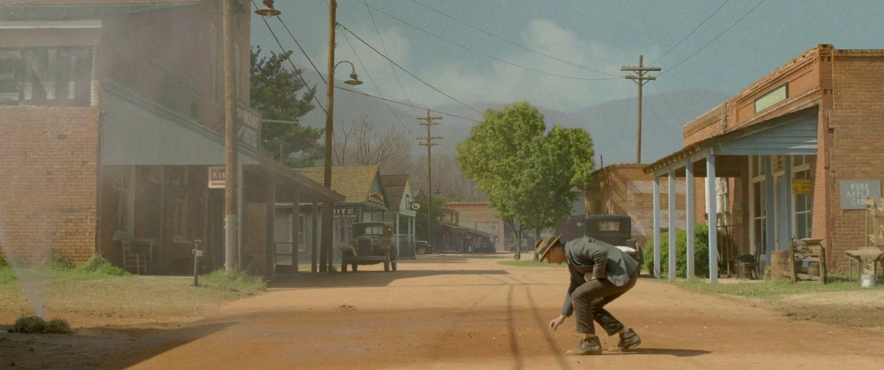 Lawless (2012) movie still: wide — A rural 1940s town with brick buildings and a dirt road under soft daylight. A man in a suit crouches in the…