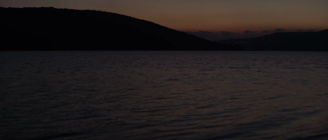 Stranger By The Lake (2013) movie still: establishing — A dark body of water sits in front of distant, silhouetted hills at sunset. The sky displays…