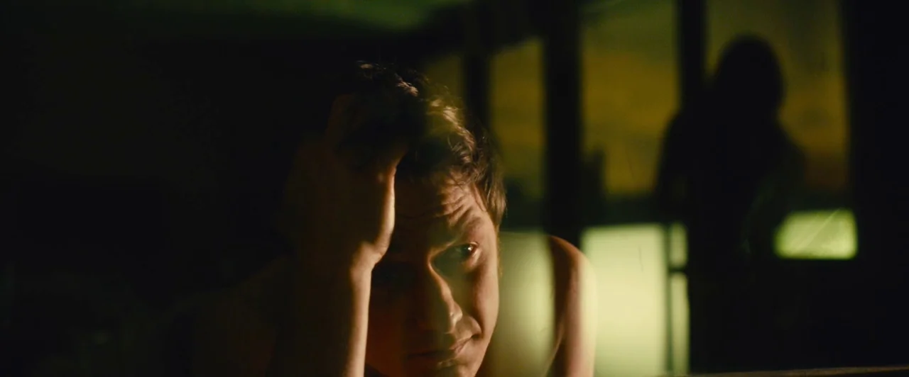 Trance (2013) movie still: closeup — A dimly lit room at night with a large window in the background. A man rests his head on his hand while…