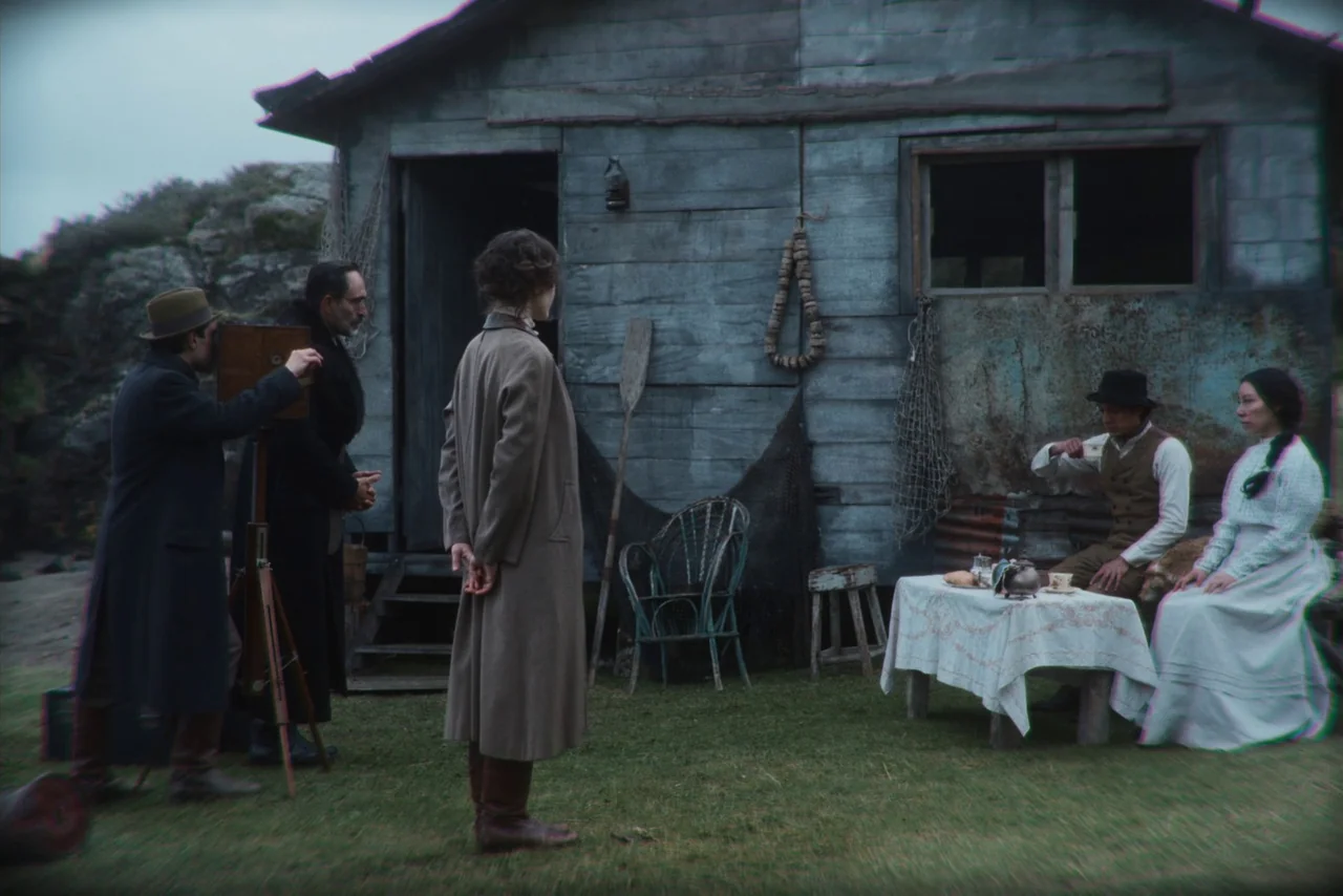The Settlers (2023) movie still: wide — A weathered wooden shack stands on a grassy patch beneath an overcast sky. A photographer sets up equipment…