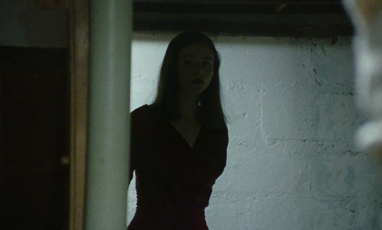 Eileen (2023) movie still: medium — A dimly lit basement features a rough, painted white cinder block wall. A woman in a dark red top stands in the…