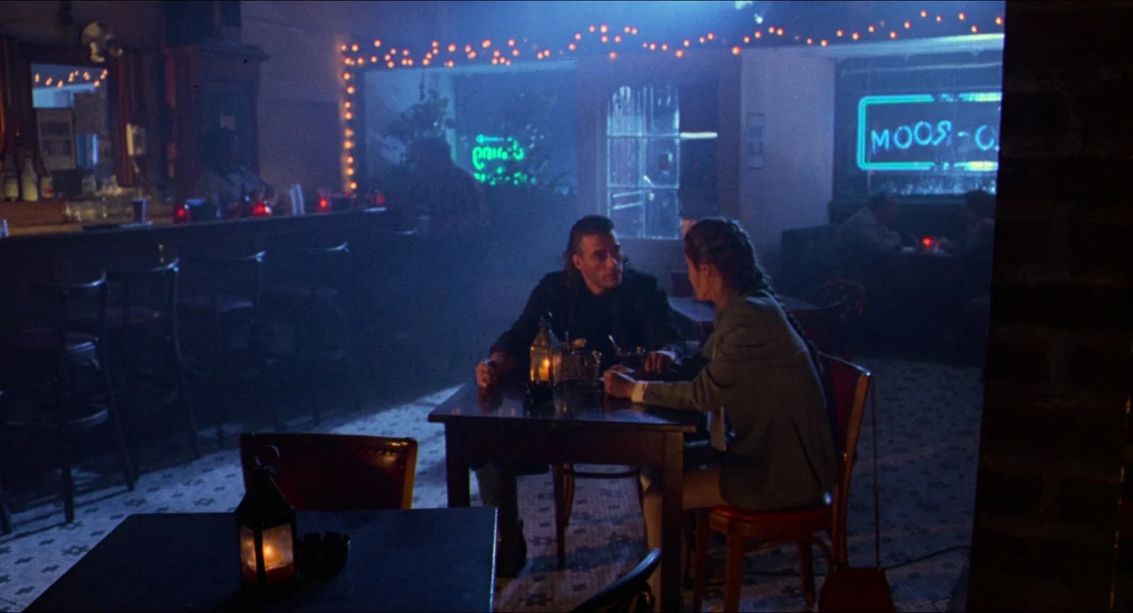 Hard Target (1993) movie still: wide — A dark, atmospheric bar features tiled floors and neon signage reflecting on the windows. A man and a woman…