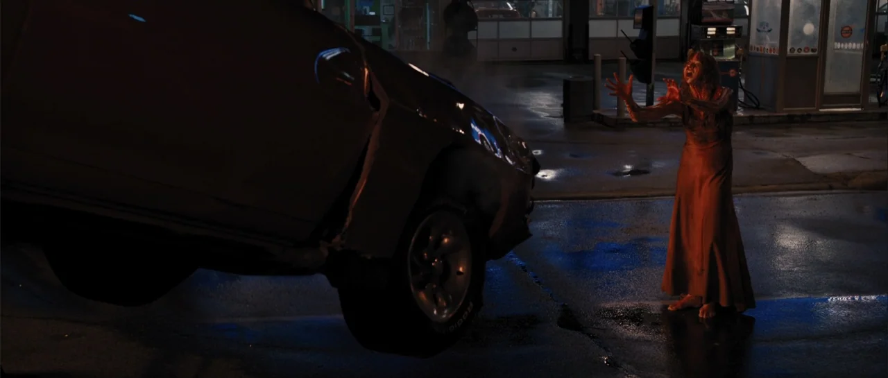 Carrie (2013) movie still: fullbody — A wet asphalt ground at a gas station at night with fuel pumps visible. A woman in a dirty dress stands with…