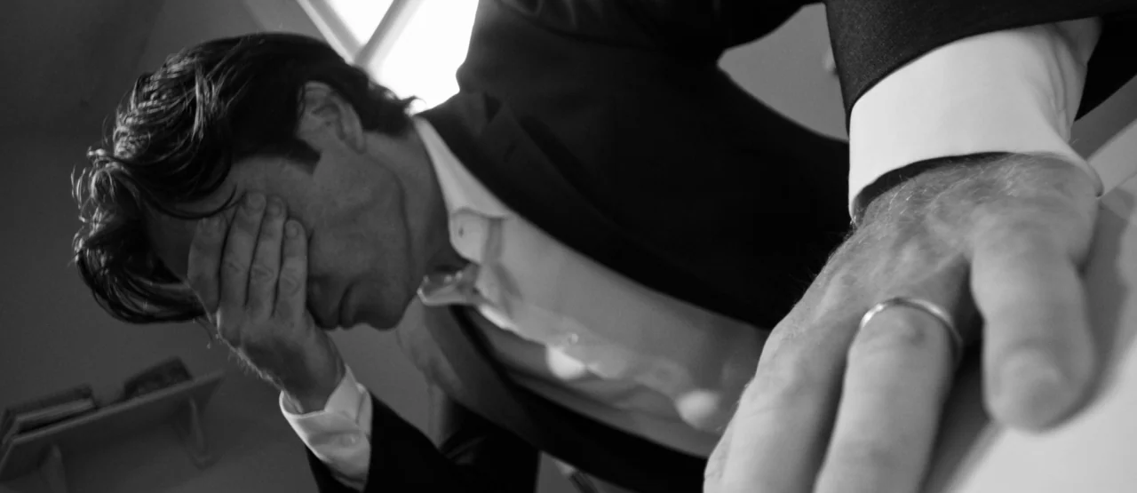 The Party (2017) movie still: closeup — A man in a suit rests his hand on his face in a dimly lit office. The camera focus highlights a wedding ring…