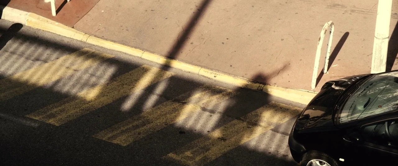 Rust And Bone (2012) movie still: establishing — An asphalt road features a yellow-striped crosswalk adjacent to a beige concrete sidewalk. A black…