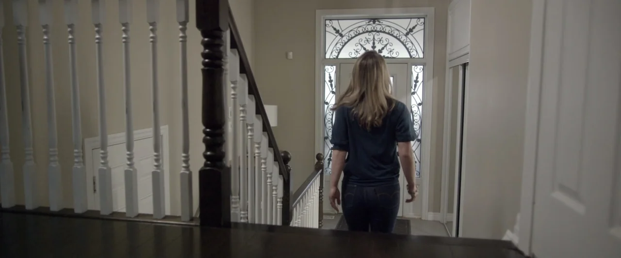 Motherly (2022) movie still: fullbody — A residential foyer features a wooden staircase with white balusters and a glass door. A woman walks away…