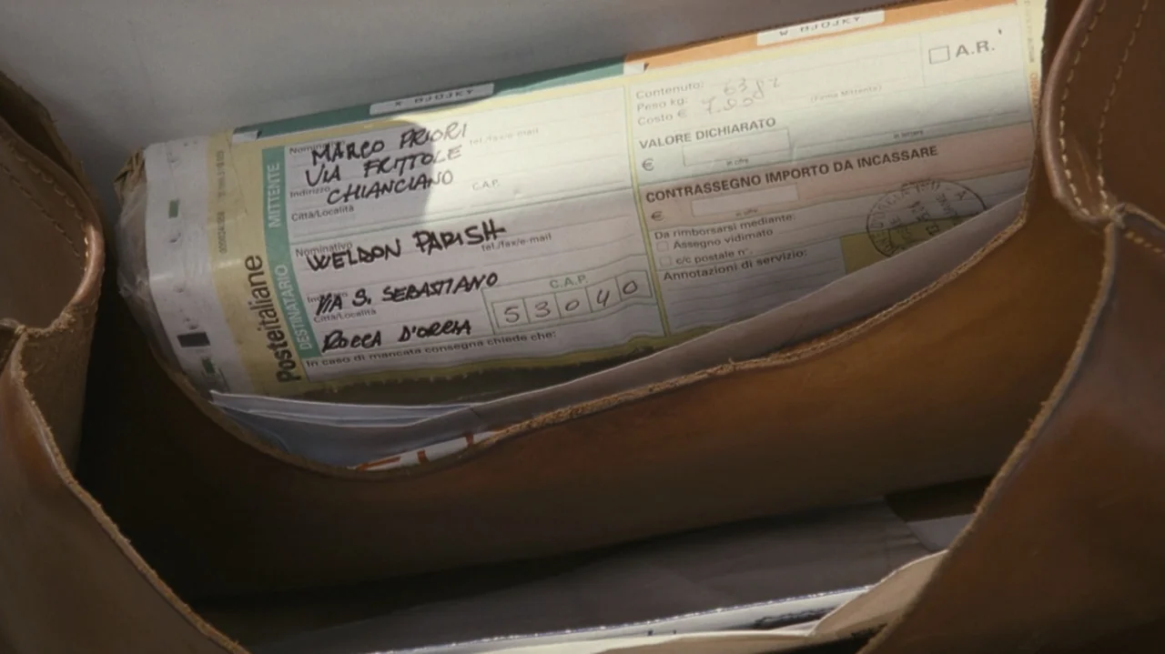 Shadows in the Sun (2005) movie still: closeup — A brown leather bag contains an Italian postal shipping form with handwritten recipient details.…