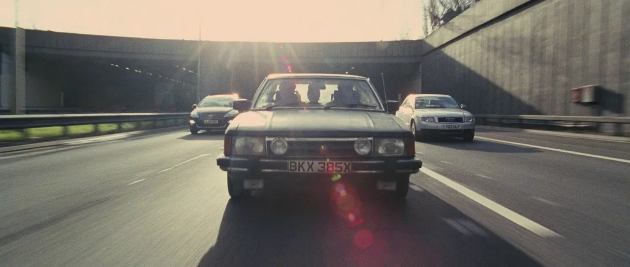 The World’s End (2013) movie still: wide — A multi-lane motorway passes under a concrete overpass during the daytime. Three cars are driving along…