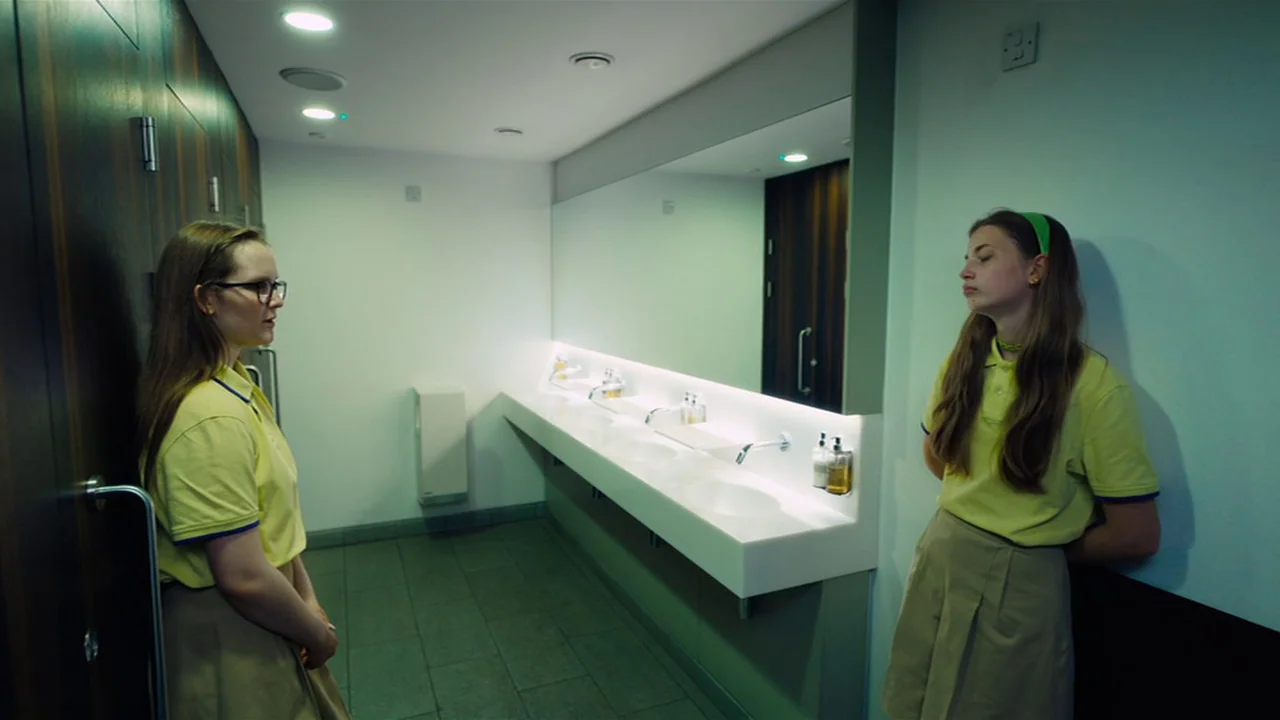 Club Zero (2023) movie still: wide — A modern, minimalist restroom features white walls, dark wood accents, and a long basin. Two girls in identical…