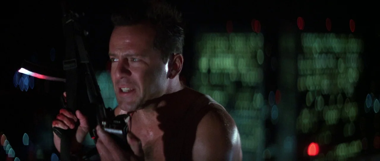 Die Hard (1988) movie still: medium — A dark night exterior high atop a building with city lights blurred in background. A sweaty man holds a…