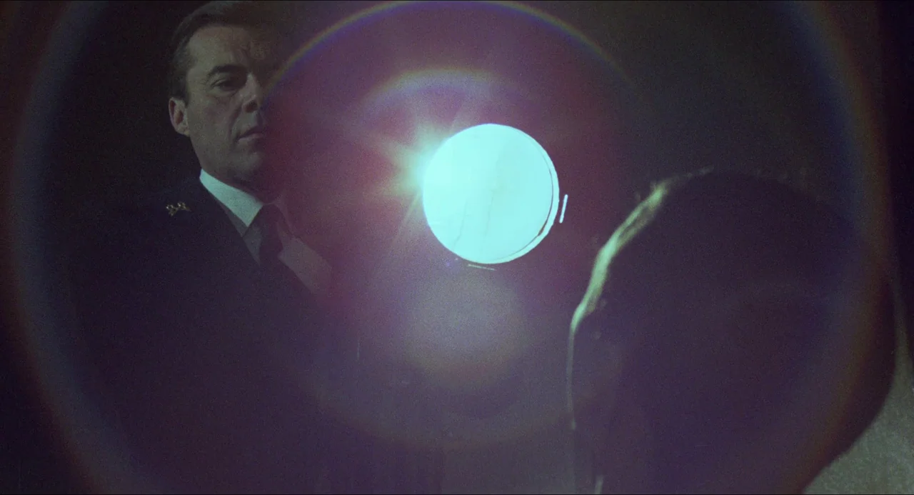 The Night Porter (1974) movie still: medium — A dark indoor setting is illuminated only by a bright circular light source. A man in a suit faces…