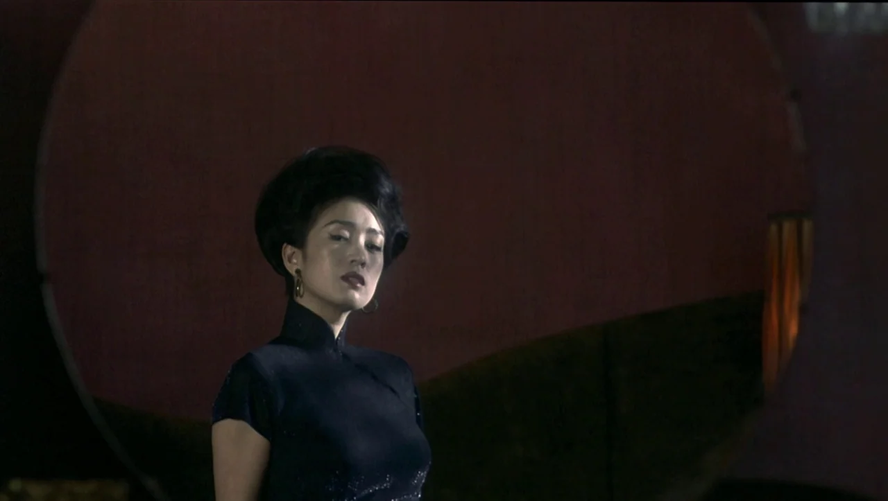 Eros (2004) movie still: medium — A woman stands in a dim room with a large circular mirror reflecting red walls. She poses against the dark…
