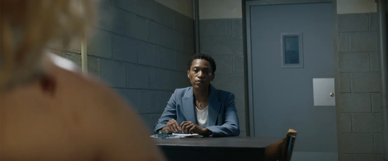 Dogman (2023) movie still: medium — An interrogation room with gray concrete block walls and a metal door. A person sits at a metal table with a…