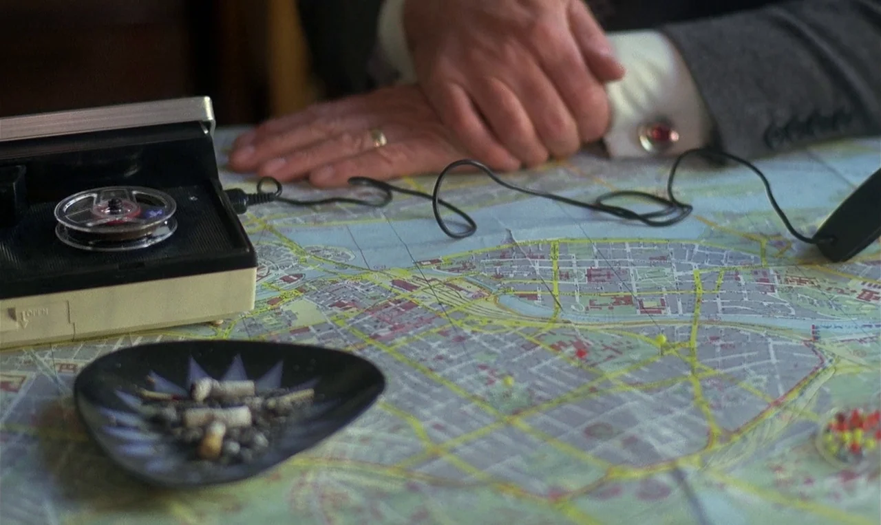 Man on the Roof (1976) movie still: closeup — A close-up view shows a detailed urban map spread across a desk surface. A tape recorder, an ashtray,…