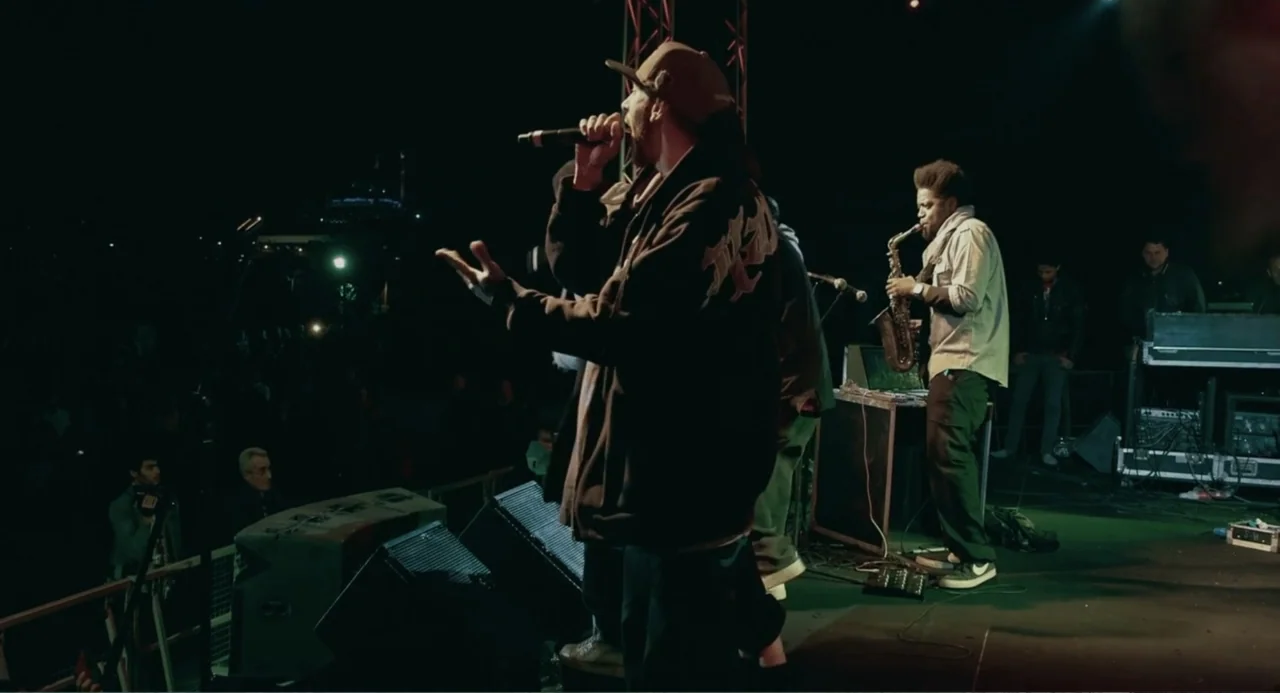 Cairo Jazzman (2017) movie still: medium — A night time music festival stage features a rapper and a saxophonist performing. The musicians occupy…