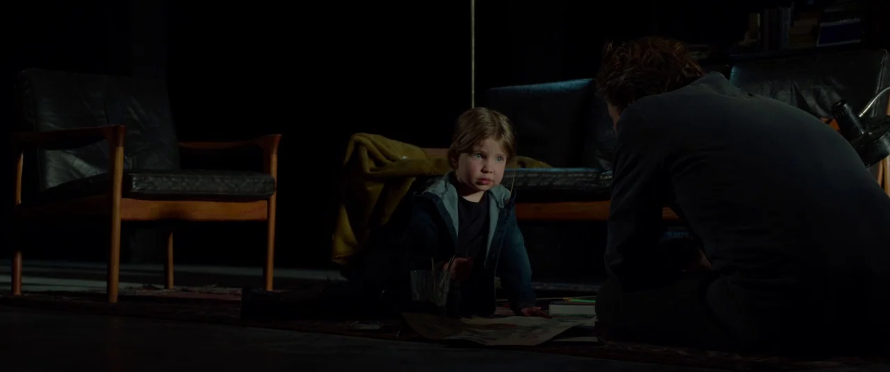 Siberia (2019) movie still: medium — A dark, minimally furnished room features two mid-century modern leather chairs on a rug. A man and a young…