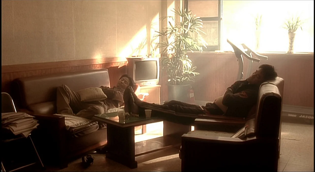 Voice (2005) movie still: wide — A sun-drenched office contains a leather sofa and chairs in a hazy atmosphere. Two men are sprawled out on the…