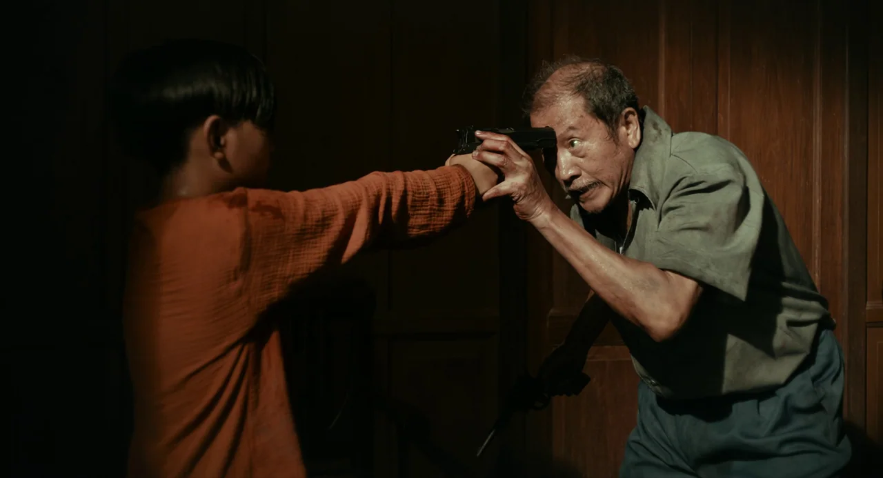 In My Mother’s Skin (2023) movie still: medium — A dark wooden interior space serves as the backdrop for a confrontation. A child holds a pistol to…