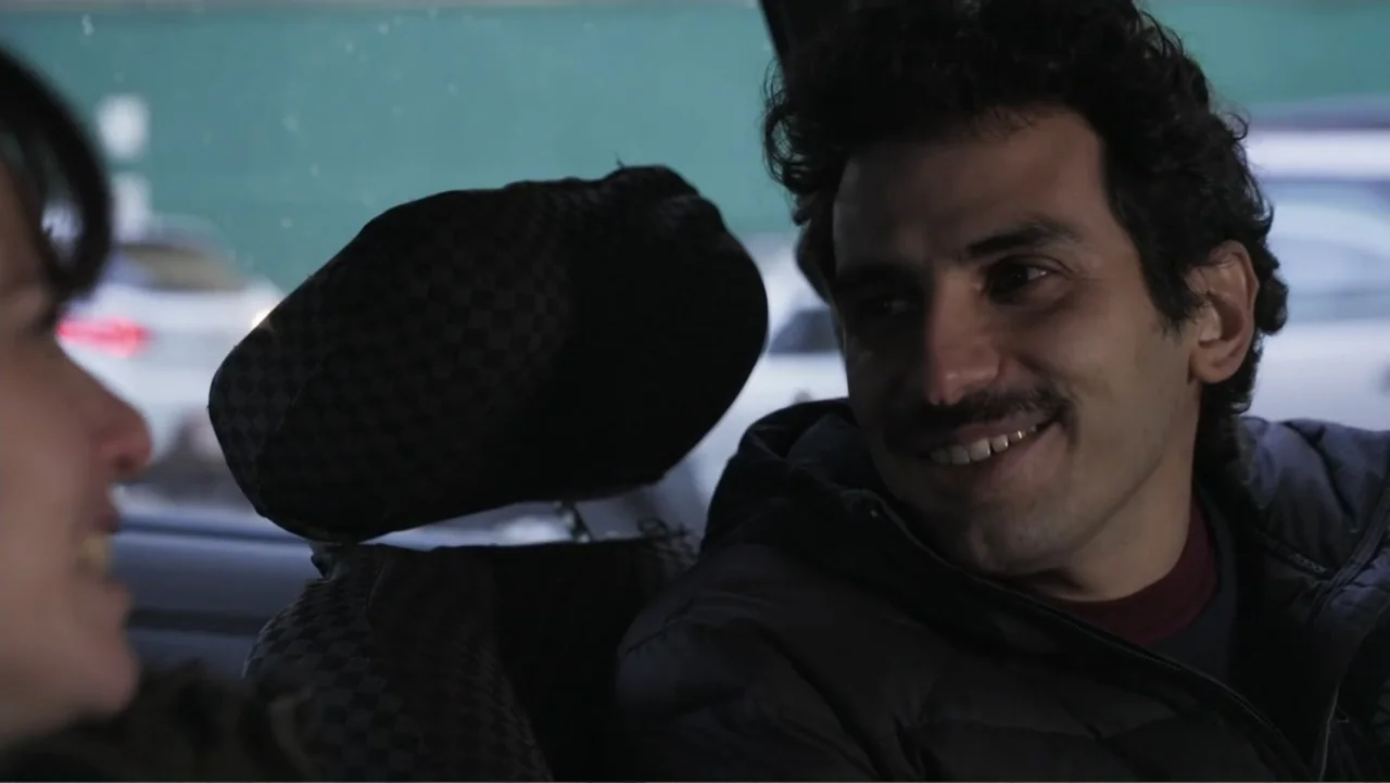 Smog in Your Heart (2022) movie still: medium — Inside a parked car during the day with a blurred urban environment outside. A man with curly hair…