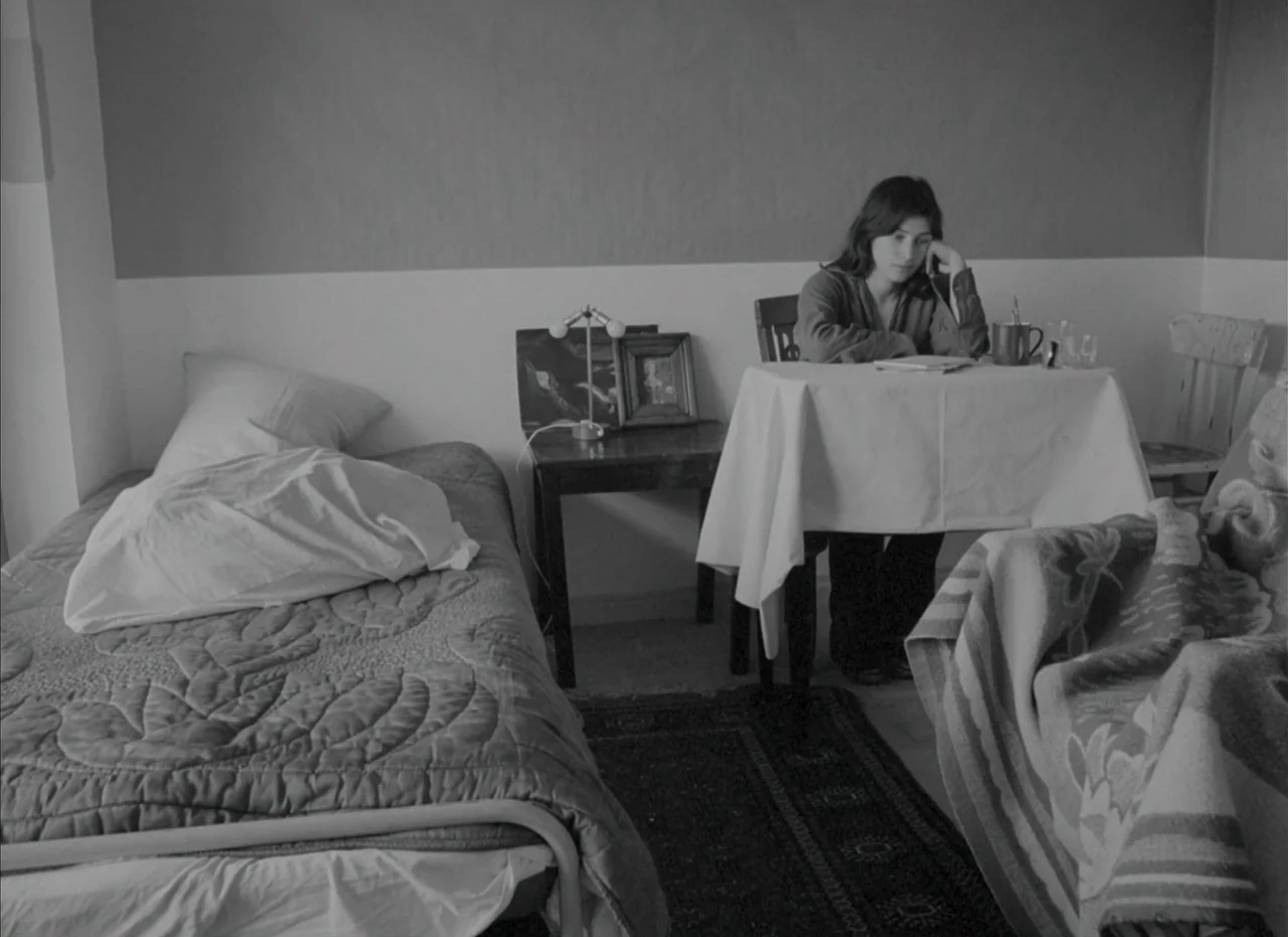 Je Tu Il Elle (1974) movie still: medium — A spare, monochromatic room features a single bed and a table with a cloth. A young woman sits at the…