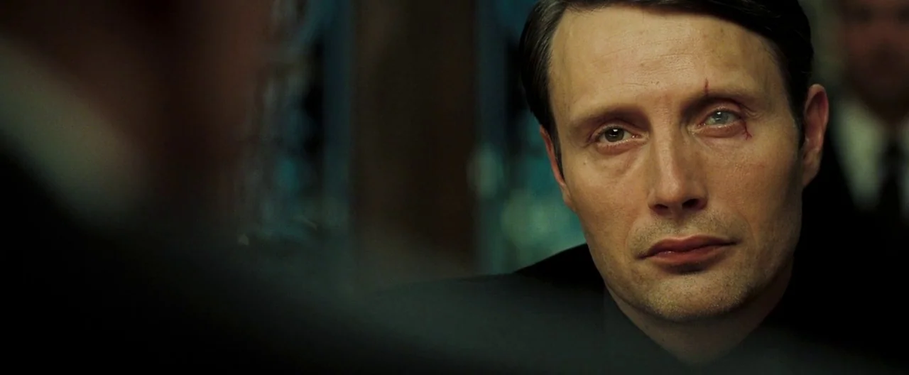 Casino Royale (2006) movie still: closeup — A dimly lit room features a man with a scarred eye staring forward. He sits across from an unseen person…